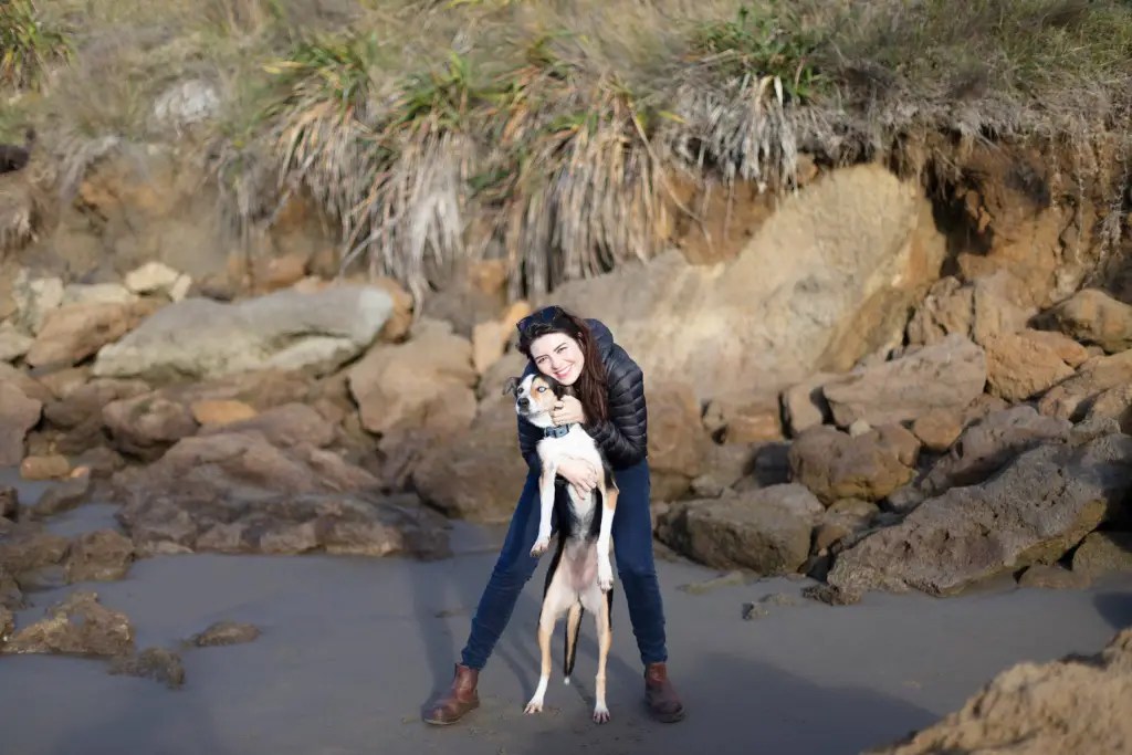 A woman hugging her dog near a rocky beach, both smiling in the soft afternoon light.