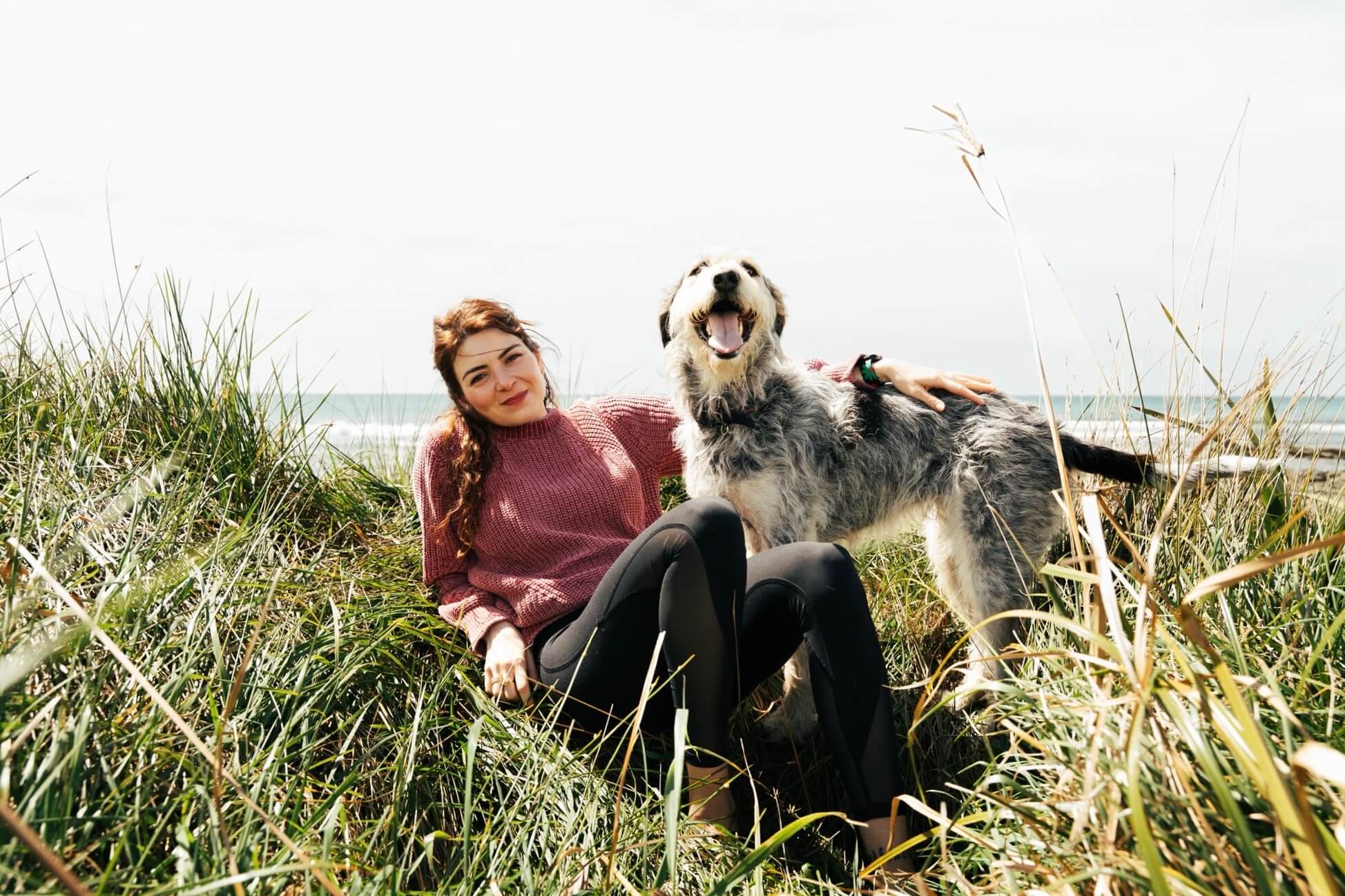 Woman relaxing on the dunes beside her smiling dog, both bathed in sunlight and surrounded by tall coastal grass.