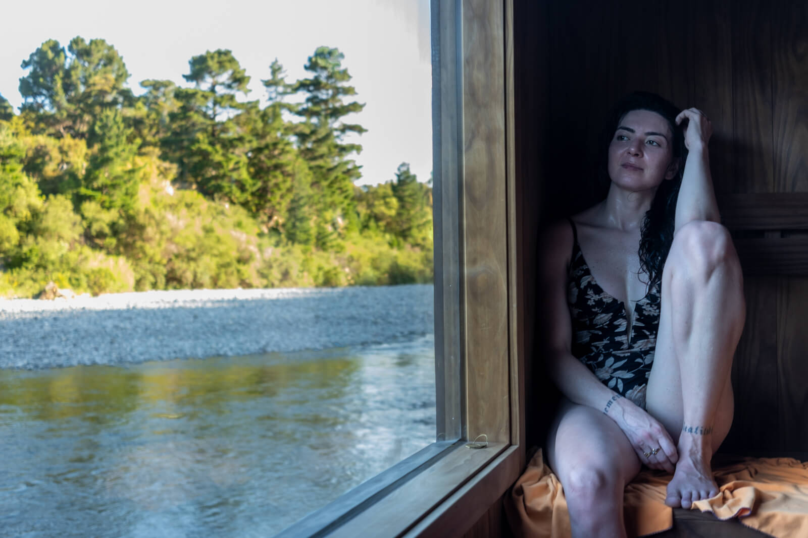 Woman sitting inside a wooden sauna by a large window overlooking a peaceful river and green forest, resting with a relaxed expression after a heat session.