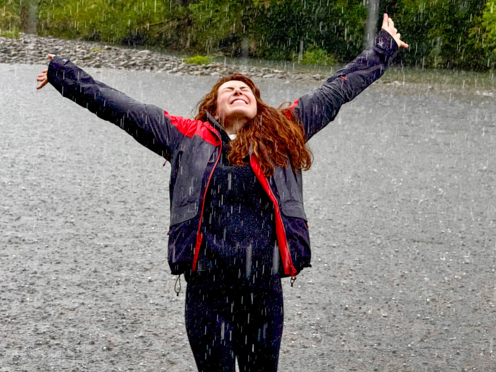 Woman standing outdoors in the rain with arms stretched wide, eyes closed, wearing a dark jacket with red details, embracing the moment in a gravel landscape surrounded by greenery.