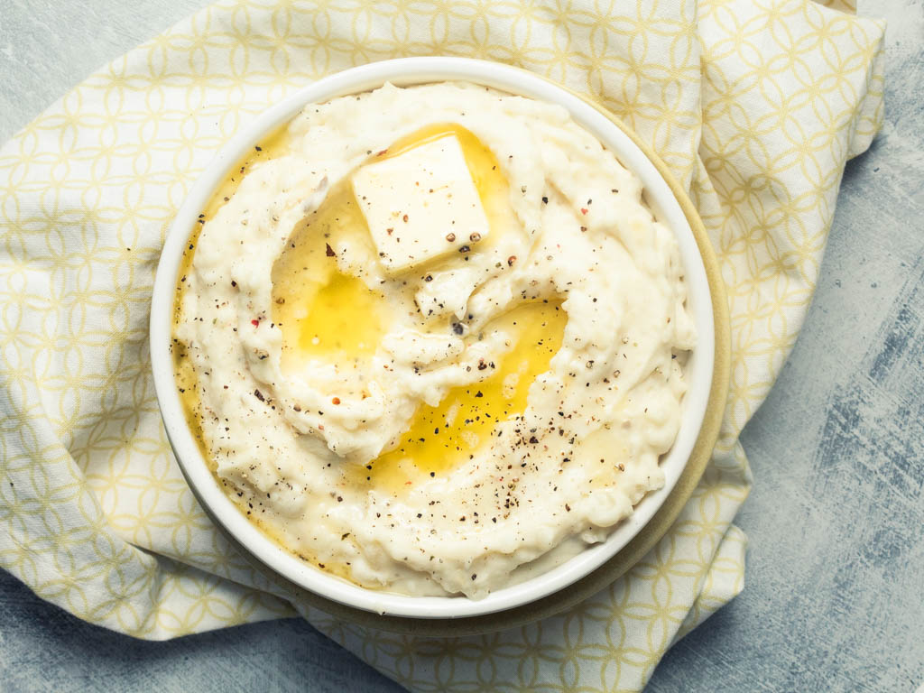 White mashed potatoes in a white bowl with yellow melted butter on top. on top of a yellow and white table napkin on a blue gray surface