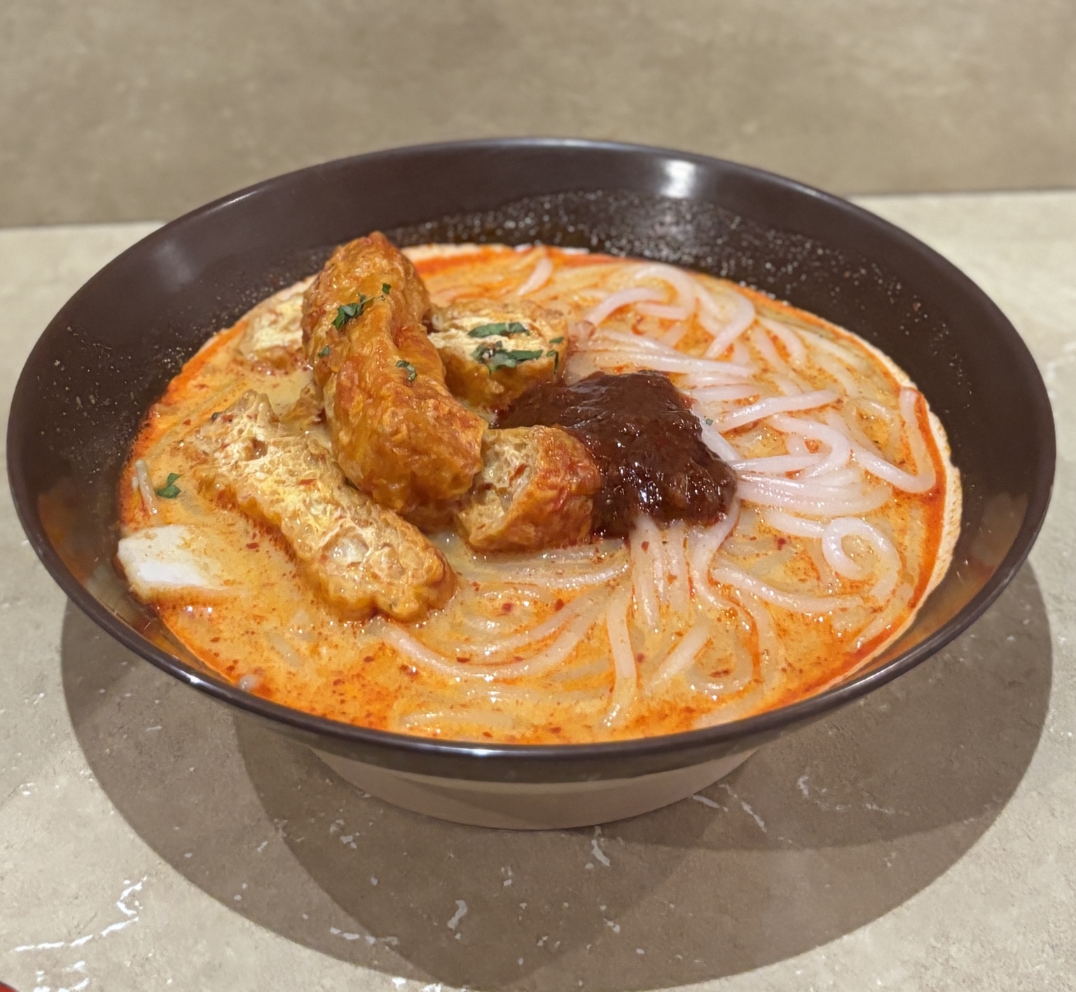 Bowl of Singapore laksa with coconut curry broth, thick rice noodles, tofu puffs, fish cake and sambal.