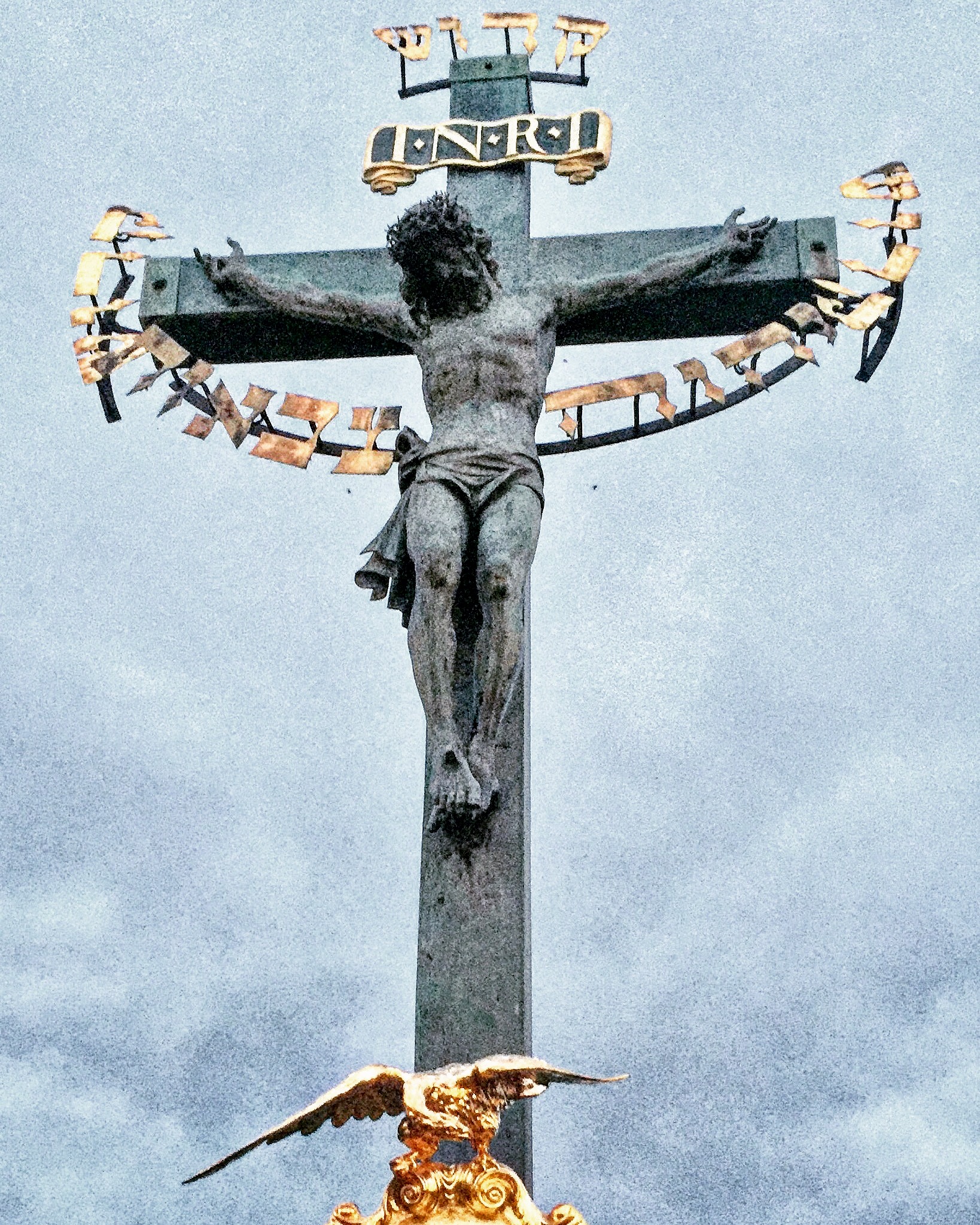 The Crucifix and Calvary (Prague). There are 30 statues mounted to the balustrade of Charles Bridge in Prague. This sculpture is one of the most historically interesting sculptures on the bridge, which gradually gained its present appearance throughout many centuries. The original wooden crucifix was installed at this place soon after 1361 and probably destroyed by the Hussites in 1419. A new crucifix with a wooden corpus was erected in 1629 but was severely damaged by the Swedes towards the end of the Thirty Years' War. The remnants of this crucifix can be found in the lapidarium of the National Museum in Prague. This was replaced by another wooden Calvary which, in turn, was replaced with a metal version in 1657. Bought in Dresden, this crucifix was originally made in 1629 by H. Hillger based upon a design by W. E. Brohn. In 1666, two lead figures were added, but these were replaced in 1861 by the present sandstone statues by Emanuel Max, portraying the Virgin Mary and John the Evangelist. The golden Hebrew text on the crucifix was added in 1696 and is a prime example of Medieval European anti-Semitism. In that year, the Prague authorities accused a local Jewish leader, one Elias Backoffen, of blasphemy. As his punishment he was ordered to raise the funds for purchasing of gold-plated Hebrew letters, placed around the head of the statue, spelling out "Holy, Holy, Holy, the Lord of Hosts," the Kedusha from the Hebrew prayer and originating in the vision from the Book of Isaiah. The inscription was a symbolic humiliation and degradation of Prague Jews, forcing them to pay for a set of golden letters referring to God and hung around the neck of the statue of Christ (information from Steven Plaut, The "Vav" from the Charles Bridge). A bronze tablet with explanatory text in Czech, English and Hebrew was mounted under the statue by the City of Prague in 2000. The tablet's placement came after an American Rabbi, Ronald Brown of Temple Beth Am in Merrick, New York was passing over the bridge and noted the possibly offensive nature of the placing of the text. Upon a direct request to the mayor, the tablet was soon placed to the side of the statue.