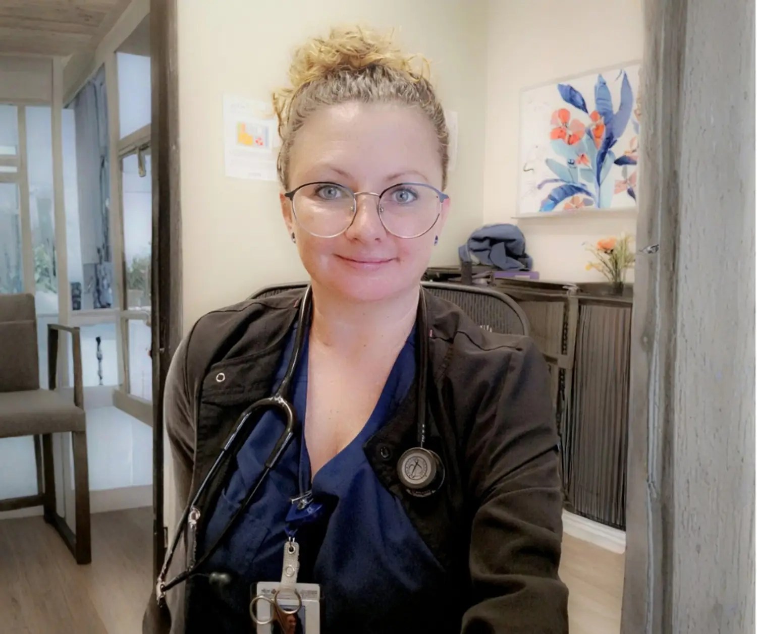 A blonde woman with glasses wearing scrubs and a stethoscope around her neck.