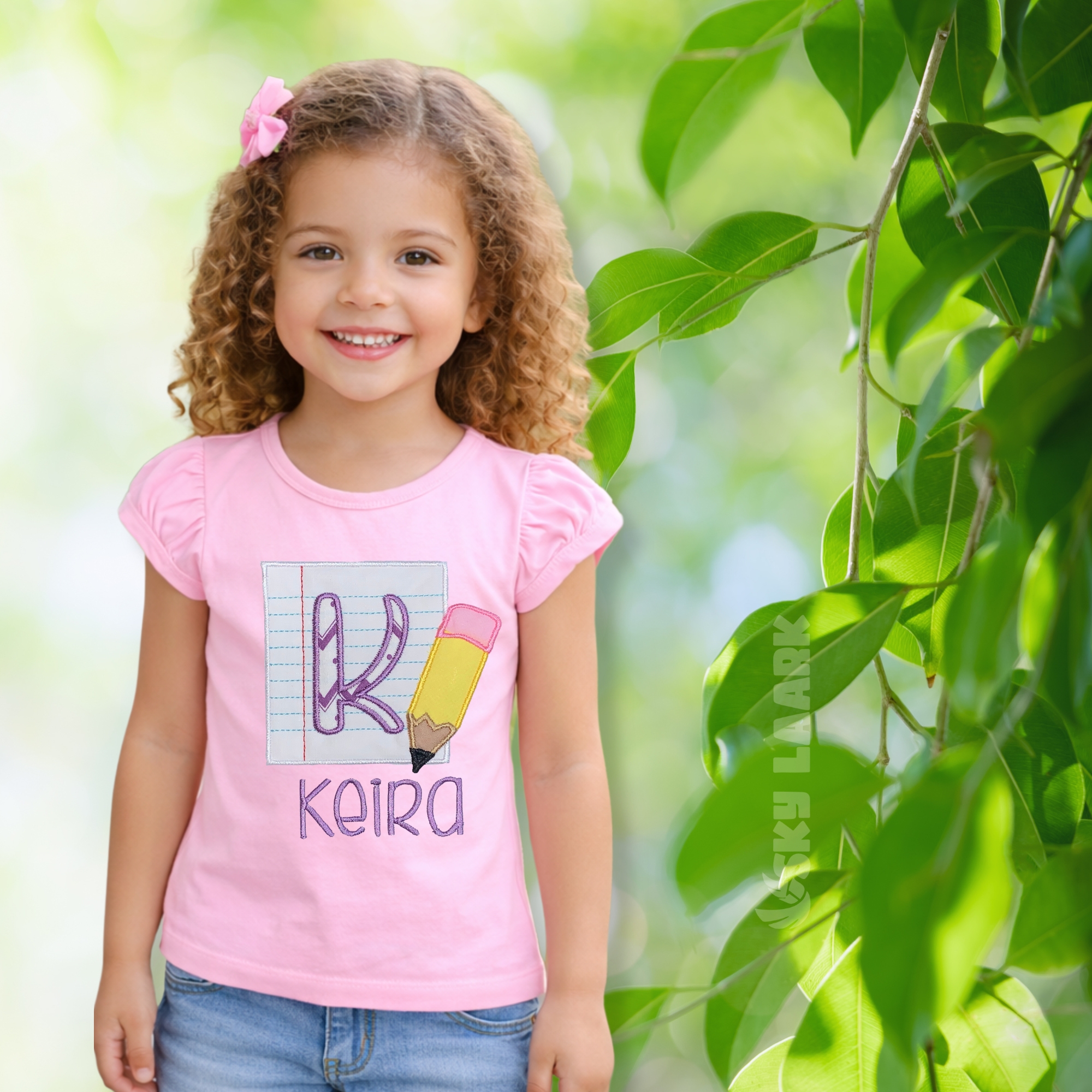 A girl wearing a personalized pink cotton back to school shirt featuring notebook paper applique, pencil embroidery, custom letter, and embroidered name on a soft 100% cotton shirt.