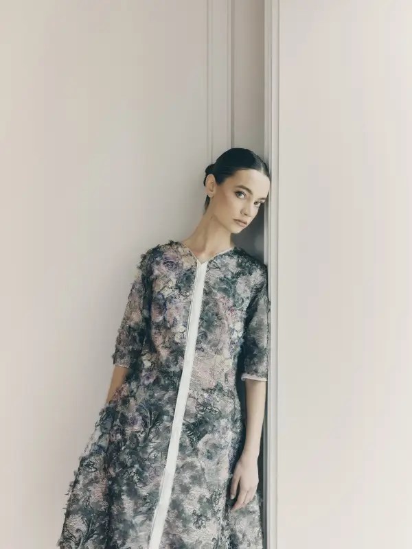 A model leans against a minimalist wall, wearing the Climbing Vine Ensemble. The intricate textile mirrors ivy weaving up the walls of a centuries-old vineyard, a balance of organic movement and structured form. She is poised, her gaze steady—a fusion of nature’s quiet persistence and modern refinement.