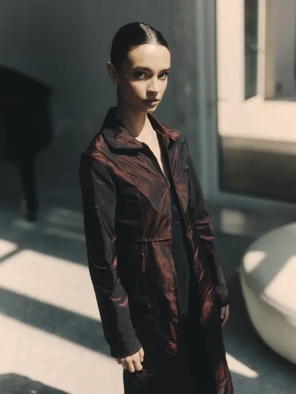 The model stands in shifting light, wearing The Umbra Coat in iridescent silk taffeta. A full-length silhouette with a subtle sheen, shifting between oxblood and deep plum. A coat built for transformation—fluid, weightless, and effortlessly adaptable.
