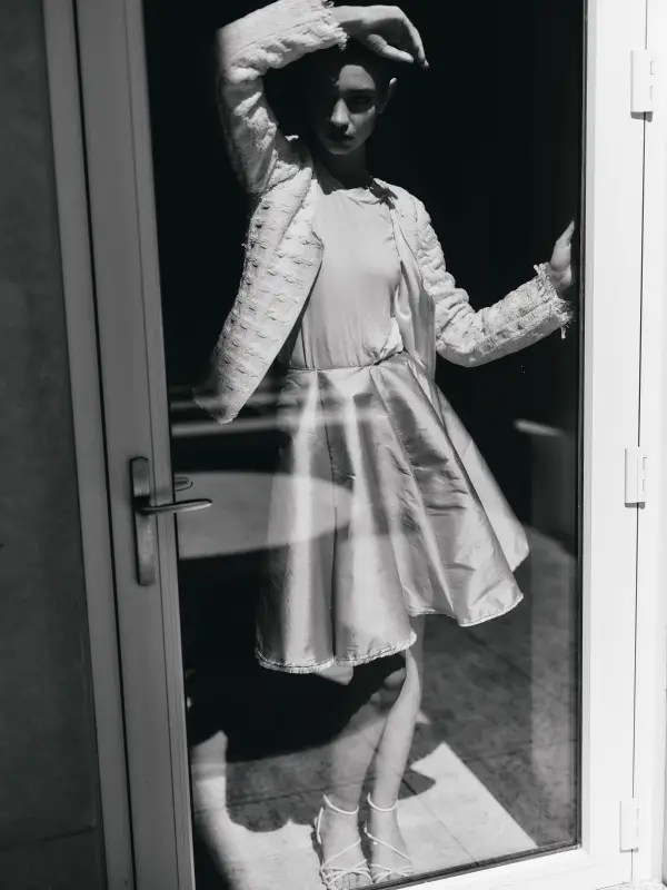 Through the circular window, a model stands at the threshold of something vast, wearing the Conque Skirt and Interplay Cardigan. The silk taffeta skirt flutters in zero gravity, its frayed ribbon hem mirroring the cardigan’s weathered sleeves—both softened by time, yet full of movement. The airlock is sealed. The cold hum of space surrounds her. Beyond the glass, a planet glows in the distance.