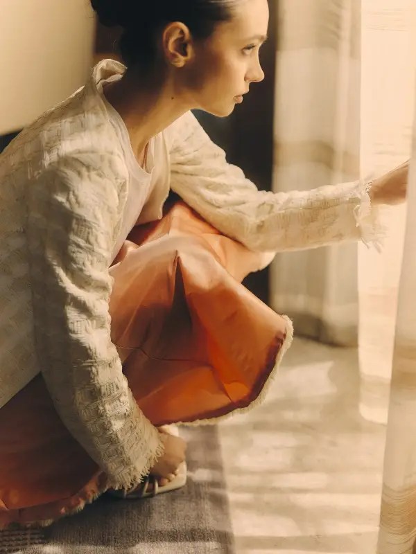 Model perched near a curtain, a model wears the Conque Skirt and Interplay Cardigan, her fingers grazing the sheer fabric as golden light filters through. The silk taffeta skirt glows—a molten ember against the soft wool texture of the cardigan. The light touches her face, her hands, the edge of the unknown.