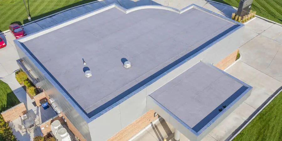 Aerial view of a commercial building featuring a low slope roofing system with a flat, gray surface. Surrounded by green grass, sidewalk, and a parking lot with red cars, the roof displays vents and distinct geometric outlines.