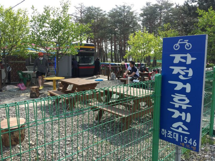 야외 나무 테이블과 의자가 마련된 자전거 휴게소에서 사람들이 휴식을 취하고 있다.