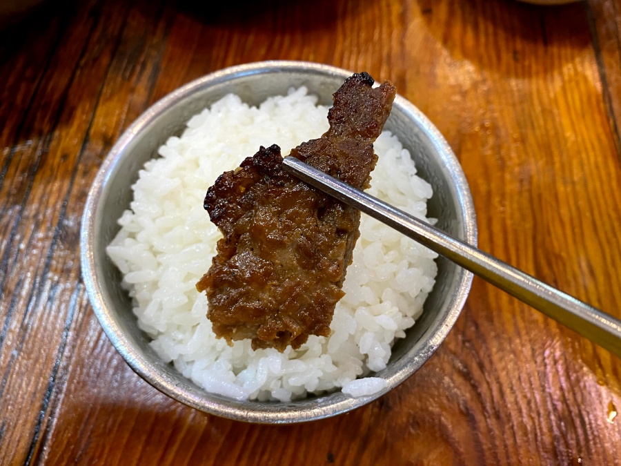 젓가락으로 집어 올린 구운 고기 한 점과 흰 쌀밥이 담긴 밥그릇