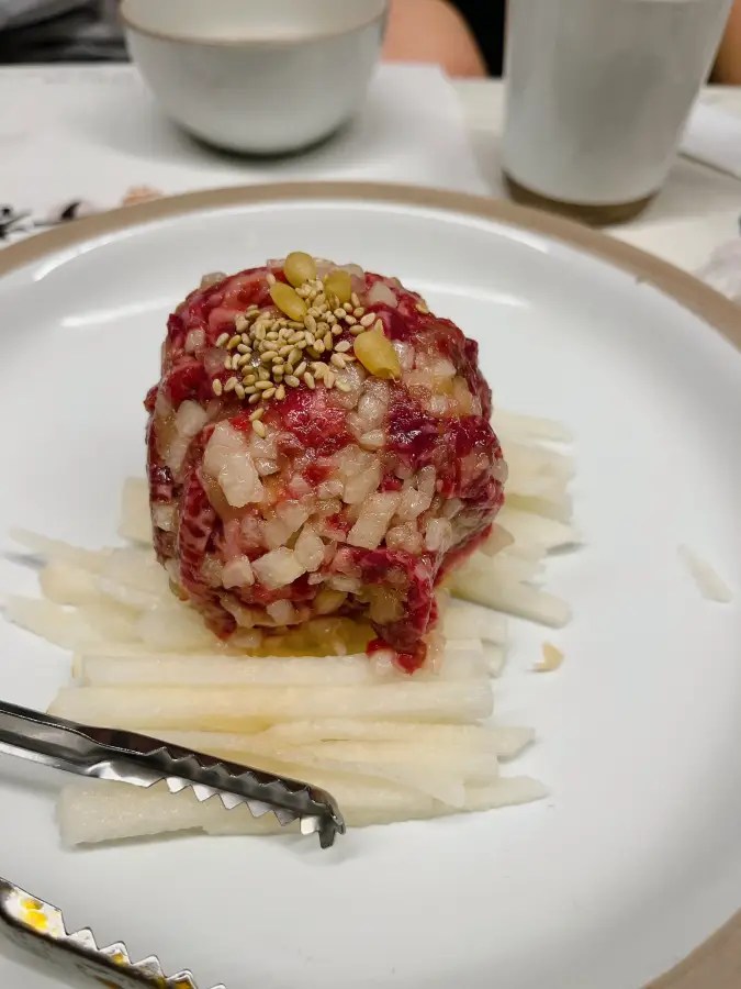 채 썬 배 위에 둥글게 뭉쳐진 육회와 그 위에 뿌려진 잣과 깨
