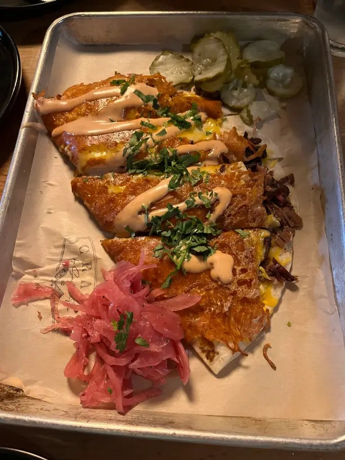 Trozos de quesadilla rociados con salsa, cebolla roja encurtida y pepinillos servidos en una bandeja forrada con papel.