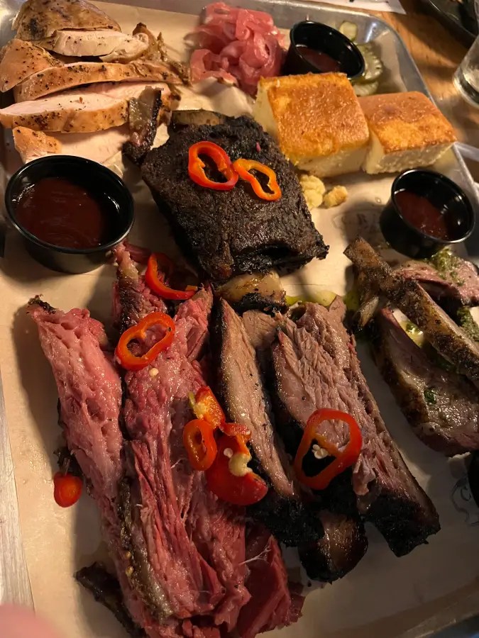 Bandeja llena de diversas carnes a la barbacoa, como brisket en rodajas finas, costillas y pollo, junto con pan de maíz.