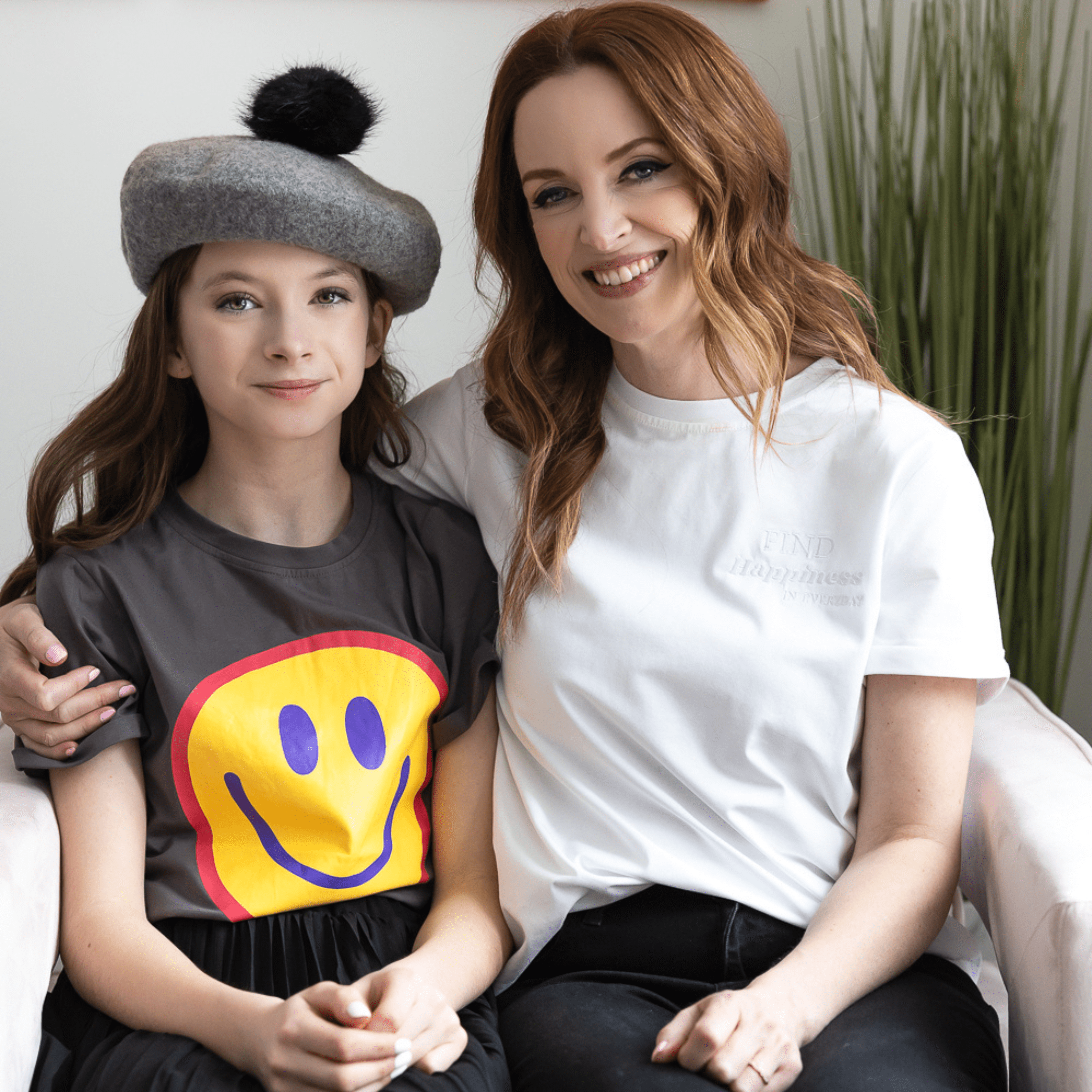 On the right, a mom with mid-length brown hair, that is curled, is sitting. She is wearing The Classic Cute Tee, smiling, and embracing her daughter. On the left, a teen girl with long, brown, curled hair is sitting. She is wearing a grey hat with a pompom on top. The teen girl is wearing the Smiley Face Tee.