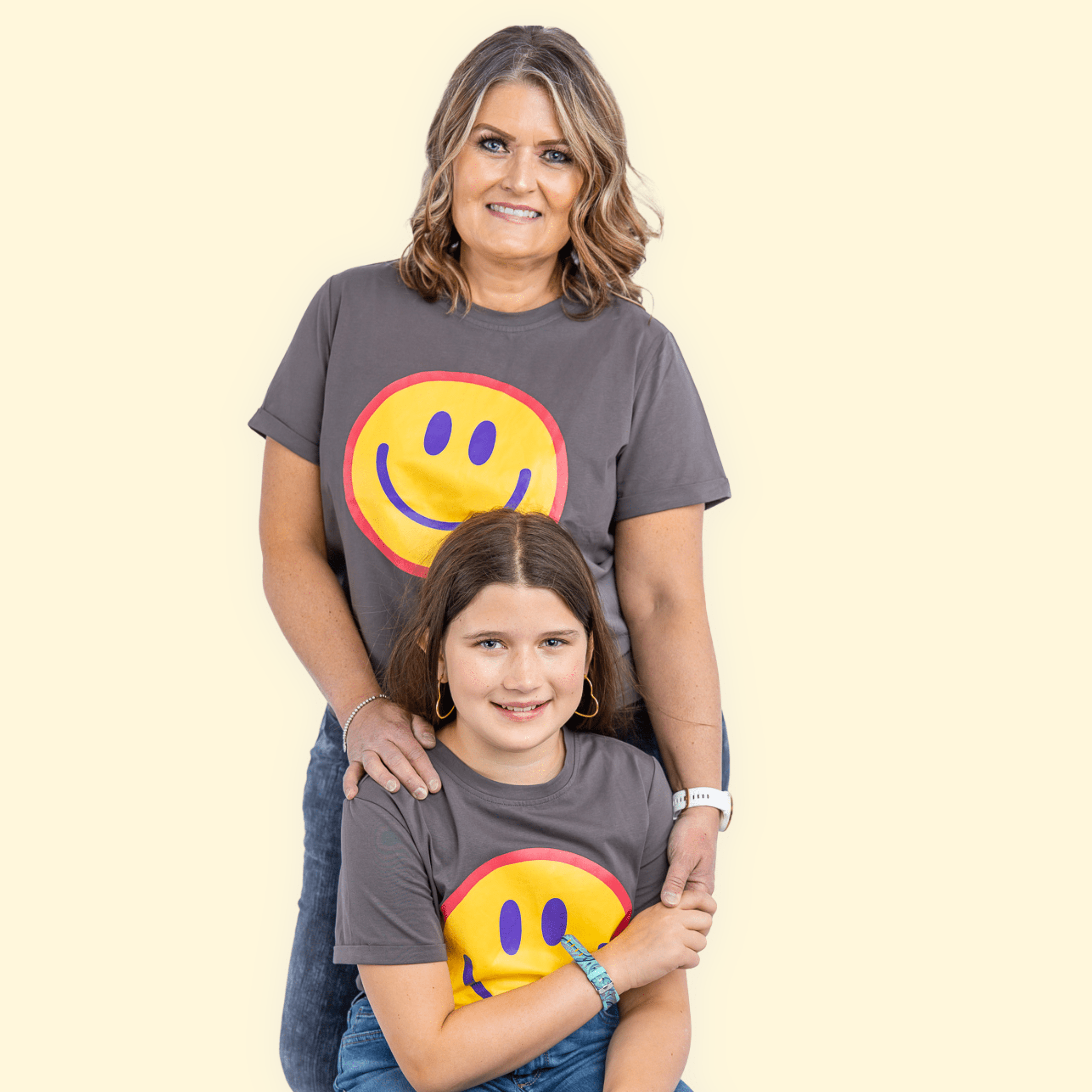 In front of a white background, a mom is standing, and her daughter is sitting on a stool. The mom has short, curled, blonde hair, and is wearing the Smiley Face Tee. The daughter has straight, short, brown hair, and is wearing the Smiley Face Tee, holding her mom's hand.