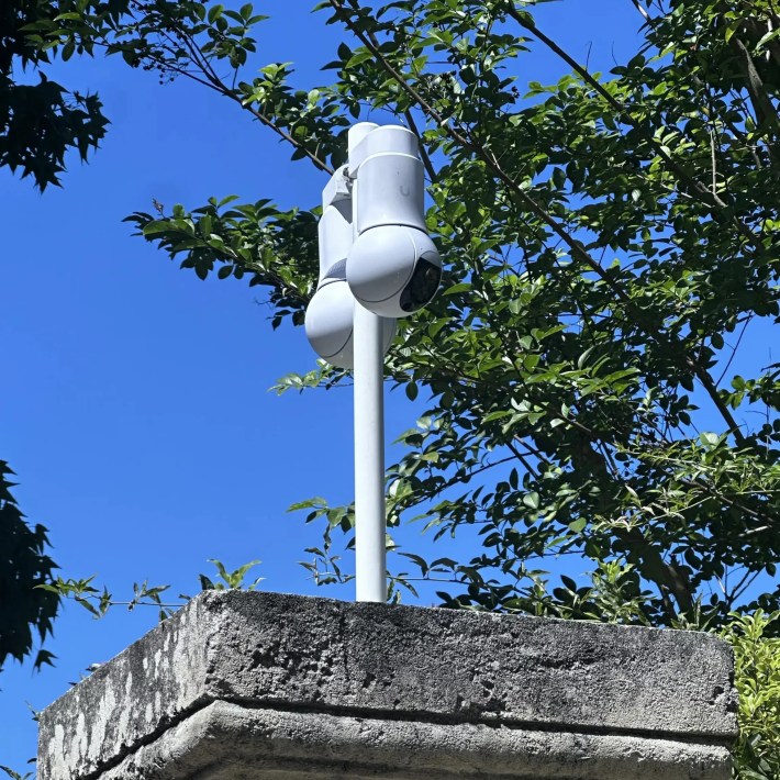 Dual security cameras mounted on a pole against a blue sky and green foliage.