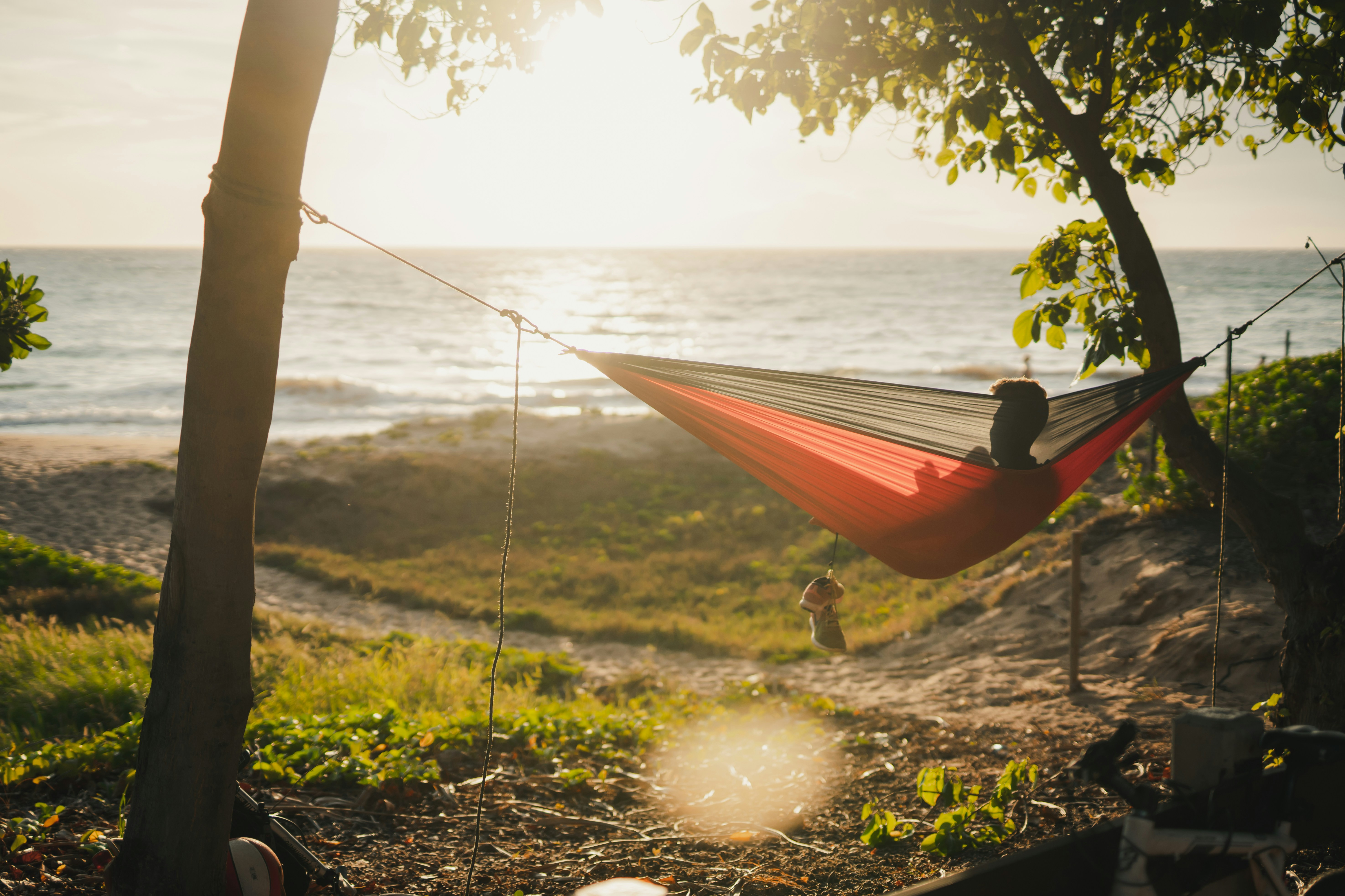 En person ligger i en röd och svart hängmatta uppspänd mellan två träd vid en sandstrand. Solen håller på att gå ner över havet, vilket skapar ett gyllene sken. Träden och lövverket ramar in scenen, och en liten väska hänger från hängmattan.