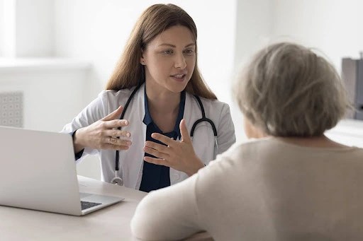 Positive doctor talking to elderly patient at office workplace, speaking, smiling with hands gestures, meeting with old lady at table with laptop, giving medical help.