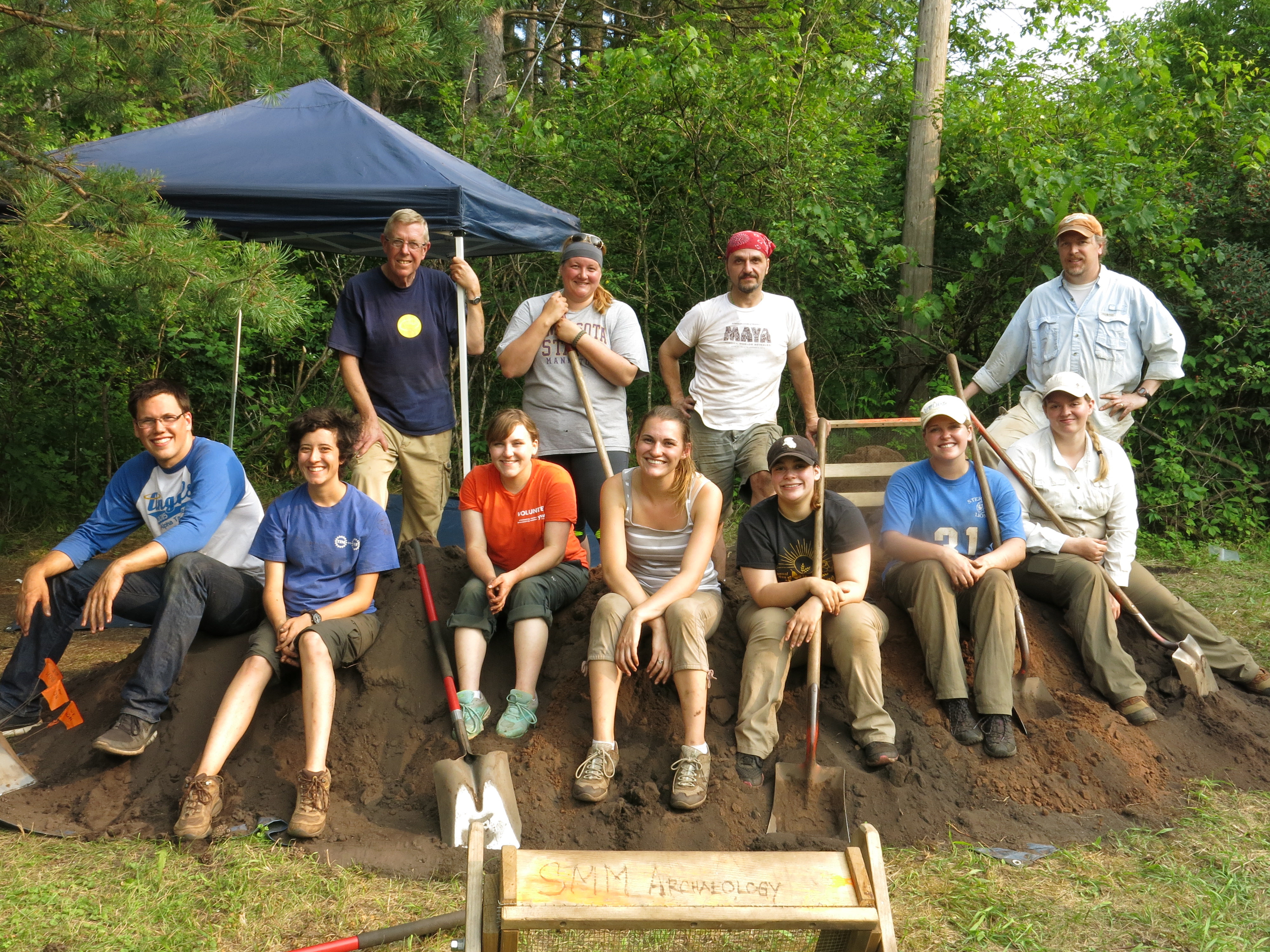 A group of students with shovels sit on a dirt mound with trees around them during Archaeology Field School with Dr. Ed Fleming.