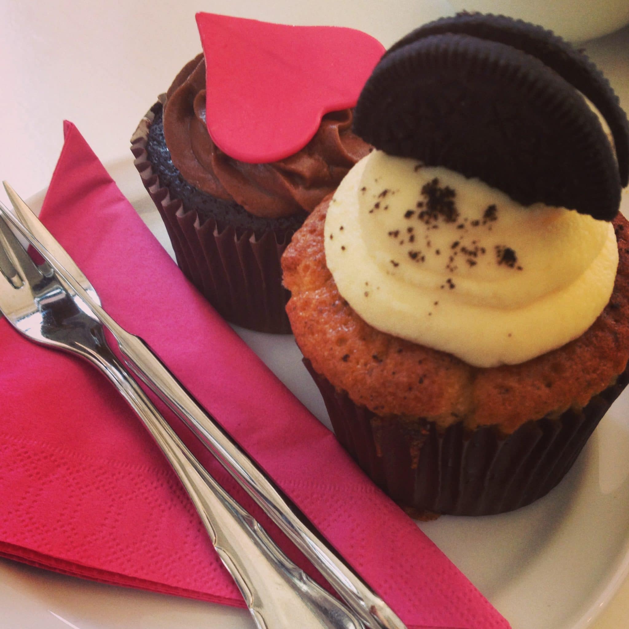 Zwei Cuperella-Cupcakes auf einem weißen Teller: einer mit Schokoladenglasur und einem roten Herz als Dekoration, der andere mit Sahneglasur und einem Schokoladenkeks obendrauf. Daneben stehen eine rosa Serviette, eine silberne Gabel und ein Messer.
