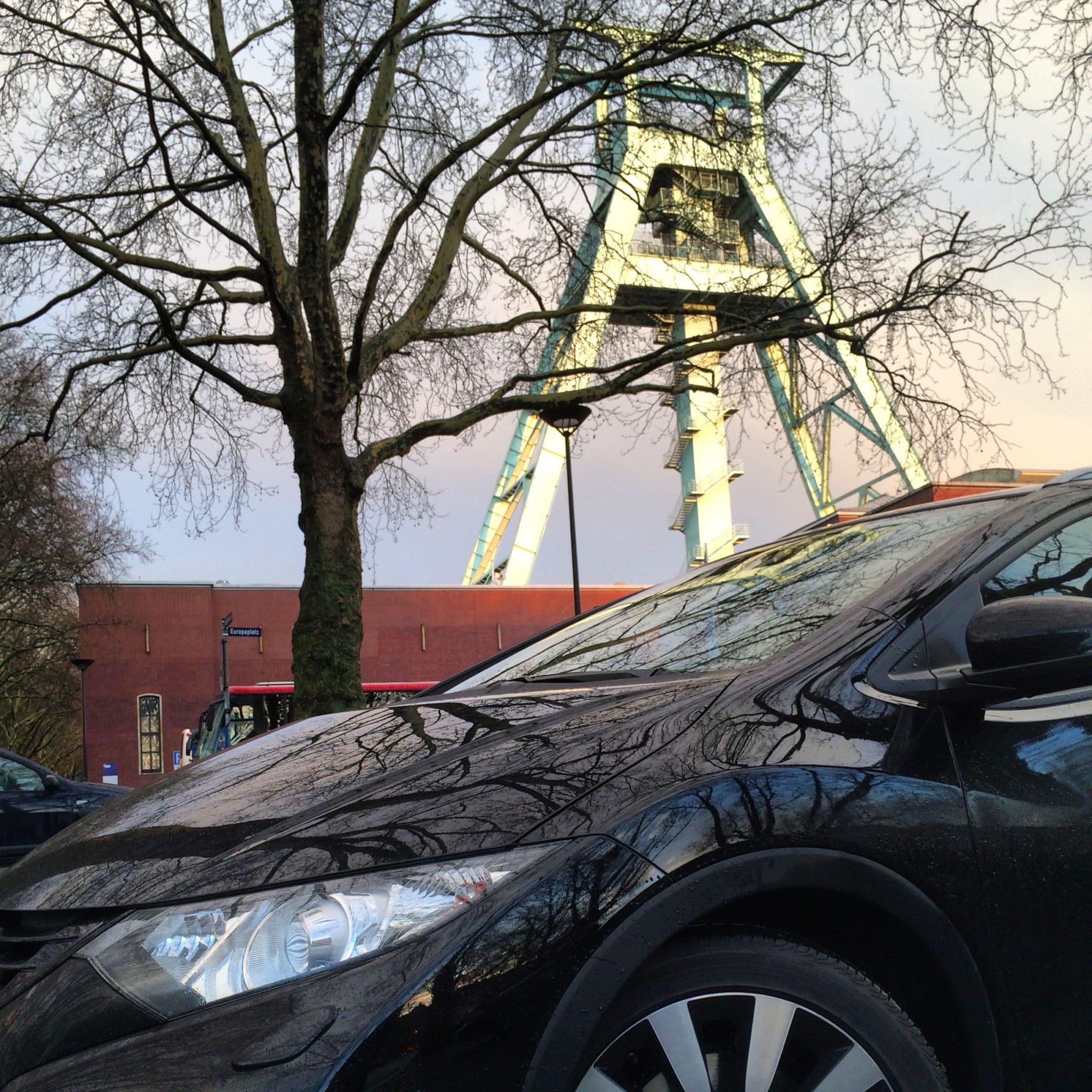 Im Vordergrund ist ein schwarzer Honda Civic geparkt, über dem kahle Äste hängen. Im Hintergrund ragt vor einem wolkigen Himmel ein hohes, grünes Industriegebäude aus dem Bergbau empor. Hinter dem Gebäude ist teilweise ein rotes Backsteingebäude zu sehen.