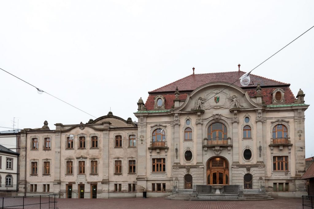Ein historisches Steingebäude in Colmar mit kunstvoller Architektur, Rundbogenfenstern, dekorativen Schnitzereien und einem roten Ziegeldach. Die Fassade weist Balkone und einen bogenförmigen Eingang auf, der auf einen gepflasterten Platz unter einem bedeckten Himmel ausgerichtet ist.