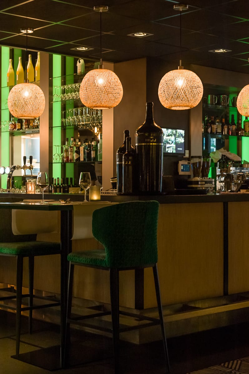 Eine gemütliche Bar in Enschede besticht durch eine gedämpfte Atmosphäre. Drei geflochtene Pendelleuchten hängen über der Theke, auf der Flaschen und Gläser ordentlich angeordnet sind. Grünes Hintergrundlicht hebt die Regale hervor, während zwei hohe Stühle mit grünem Bezug die Gäste zum Entspannen an der Bar einladen.