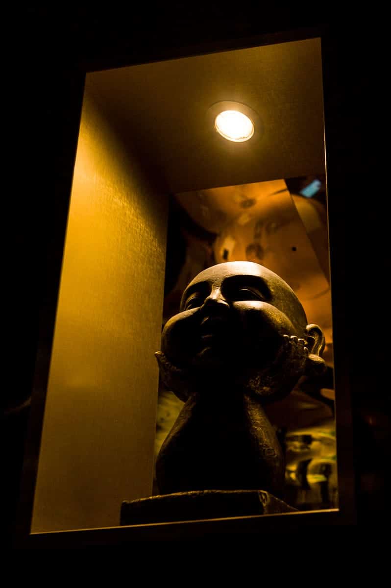 In einer schwach beleuchteten Nische wird eine Büste mit geschlossenen Augen und Händen unter dem Kinn kunstvoll zur Schau gestellt. Das sanfte Deckenlicht wirft dramatische Schatten auf die strukturierte Oberfläche, alles vor einem warmen goldenen Hintergrund, der an die Dämmerungsfarben von Enschede erinnert.