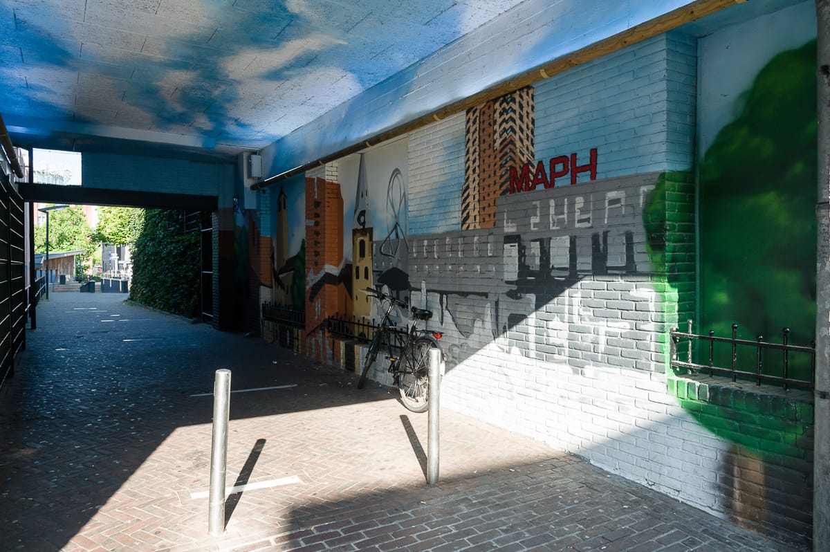 Ein Wandgemälde in einer Gasse in Enschede zeigt urbane Szenen, darunter Gebäude, Bäume und eine Brücke. An der bemalten Wand lehnt ein Fahrrad. Sonnenlicht wirft geometrische Schatten auf den Ziegelweg, die einen Kontrast zum schattigen Bereich bilden. An der Decke ist ein blauer Himmel mit Wolken zu sehen.