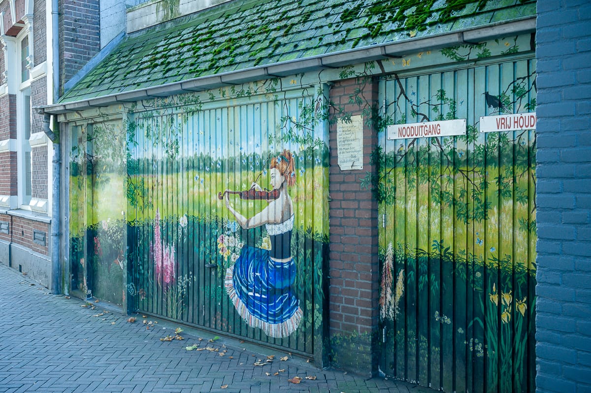 Ein Wandbild an einem Garagentor in Enschede zeigt eine Frau in einem blau-weißen Kleid, die auf einer Wiese Geige spielt. Der üppige Hintergrund zeigt grüne Felder und Bäume, darüber ein moosbedecktes Dach. Auf beiden Seiten des Wandbilds stehen Schilder mit den Worten „NOODUITGANG“ und „VRIJ HOUDEN“.