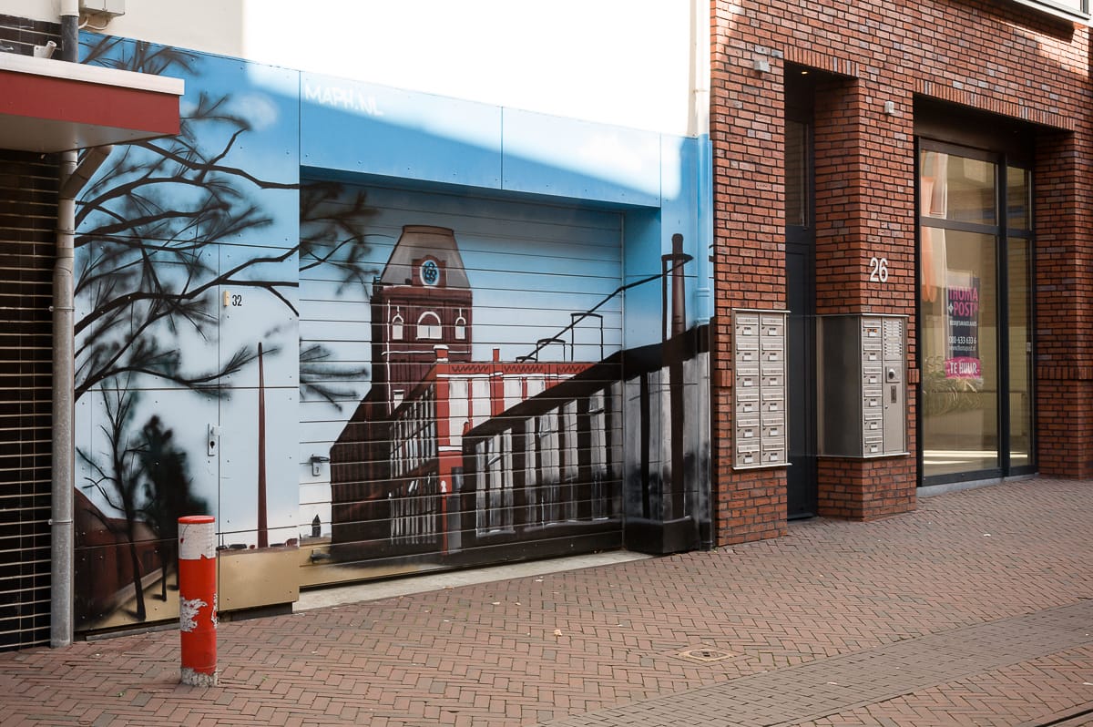Straßenkunst auf einem Rolltor in Enschede zeigt eine malerische Aussicht auf ein rotes Backsteingebäude mit einem Glockenturm, eingerahmt von dunklen Baumsilhouetten. Die Szene erstreckt sich auf angrenzende Wände. In der Nähe befindet sich eine Reihe von Briefkästen und ein weiteres Gebäude mit demselben charmanten roten Backstein und der Nummer 26 auf der Tür.