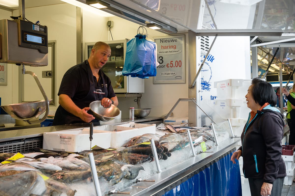 Im geschäftigen Enschede hält ein Fischhändler in schwarzer Schürze eine Metallschüssel in der Hand und unterhält sich mit einem Kunden am lebhaften Marktstand. Verschiedene Fische und Meeresfrüchte liegen auf Eis, „Nieuwe haring“ wird für 5 Euro angeboten. Die lebhafte Szene ist erfüllt von bunten Hängetaschen und Markttreiben im Hintergrund.
