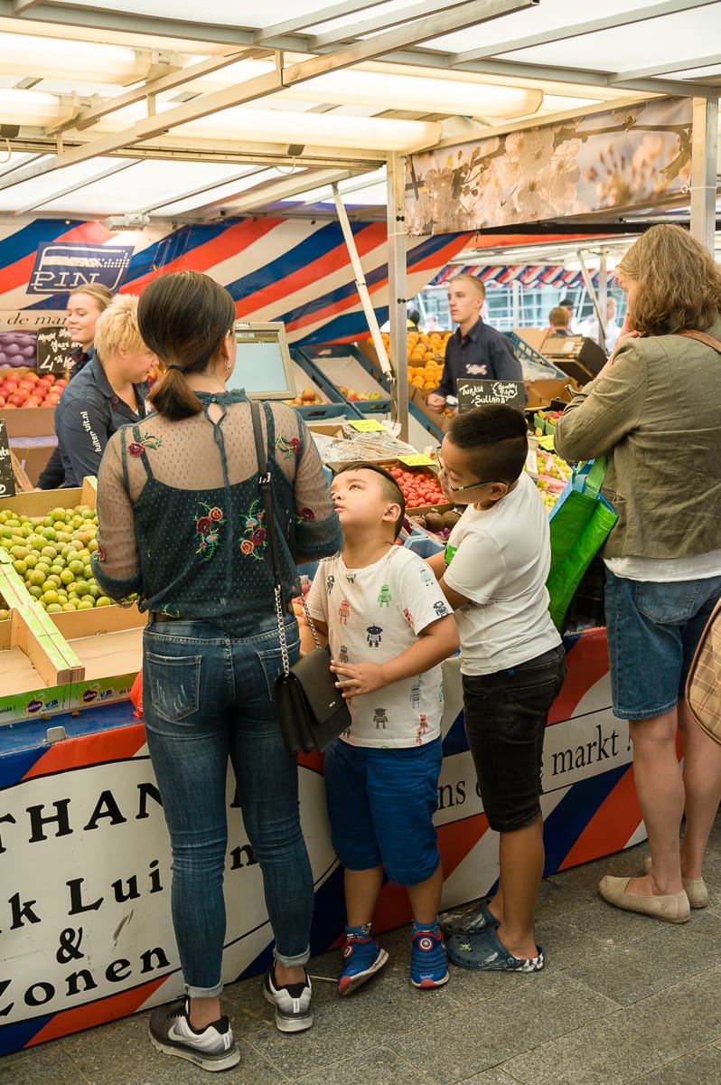 Eine Frau und zwei Kinder stehen vor einem Obststand auf dem lebhaften Markt in Enschede. Die Kinder, eines in einem weißen Hemd, das andere in einem grünen, bestaunen die bunten Früchte. Währenddessen hilft der Verkäufer in Schwarz einem anderen Kunden unter der gestreiften Plane, die ihre lebhafte Auslage abschirmt.