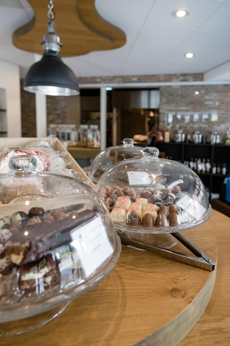 In einem gemütlichen Café in Enschede stehen auf Holztischen unter klaren Glaskuppeln verschiedene Schokoladen- und Gebäcksorten. Darüber hängt eine große schwarze Pendelleuchte. Backsteinwände, Regale mit Produkten und sanfte Beleuchtung im Hintergrund schaffen eine warme und einladende Atmosphäre.
