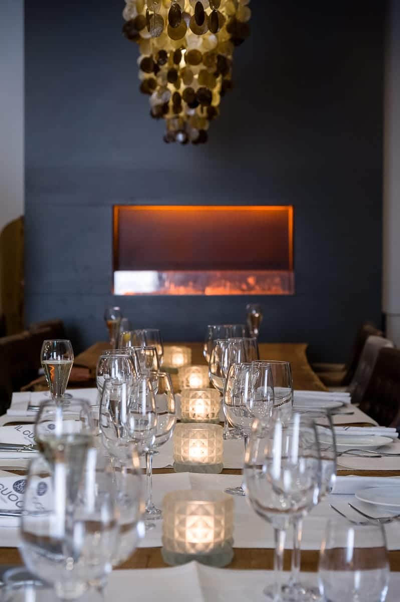Ein langer Esstisch ist elegant mit Weingläsern und Servietten gedeckt und spiegelt den kultivierten Charme von Enschede wider. Kleine brennende Kerzen säumen die Mitte. Im Hintergrund ist ein warmer, moderner Kamin in eine dunkelgraue Wand eingelassen. Darüber hängt ein einzigartiger, runder Kronleuchter, der für eine gemütliche Atmosphäre sorgt.