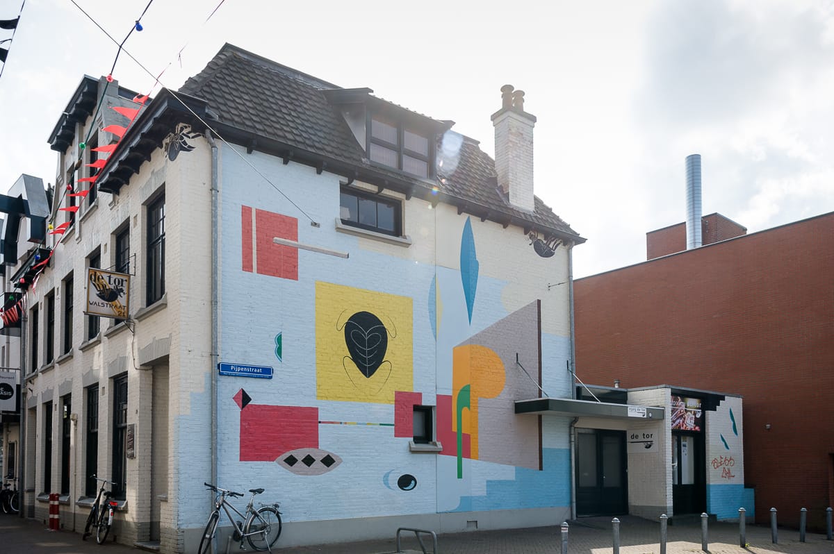 Ein zweistöckiges Gebäude in Enschede zeigt ein lebendiges, geometrisches Wandbild mit abstrakten Formen und leuchtenden Farben wie Rot, Gelb und Blau. In der Nähe des Straßenschildes sind Fahrräder geparkt. Der Himmel ist teilweise bewölkt und ein angrenzendes modernes Backsteingebäude vervollständigt die Szene.