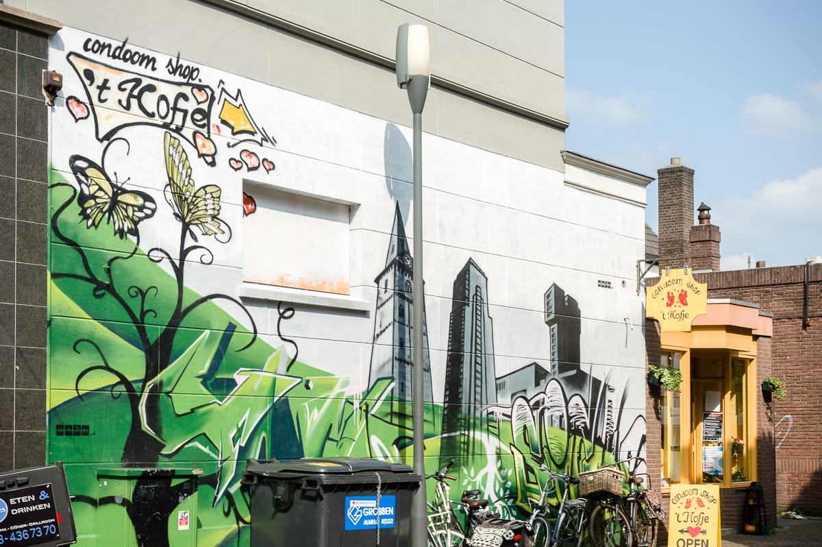 Street Art in Enschede ziert eine Hauswand mit farbenfrohen Graffitis, die Schmetterlinge, urbane Stadtlandschaftselemente und abstrakte Designs zeigen. In der Nähe hängt ein gelbes Ladenlokal mit dem Schild „Condomerie Het Hofje“ und Schildern mit den Aufschriften „geöffnet“ und „kostenlose Informationen“. Davor stehen Fahrräder.