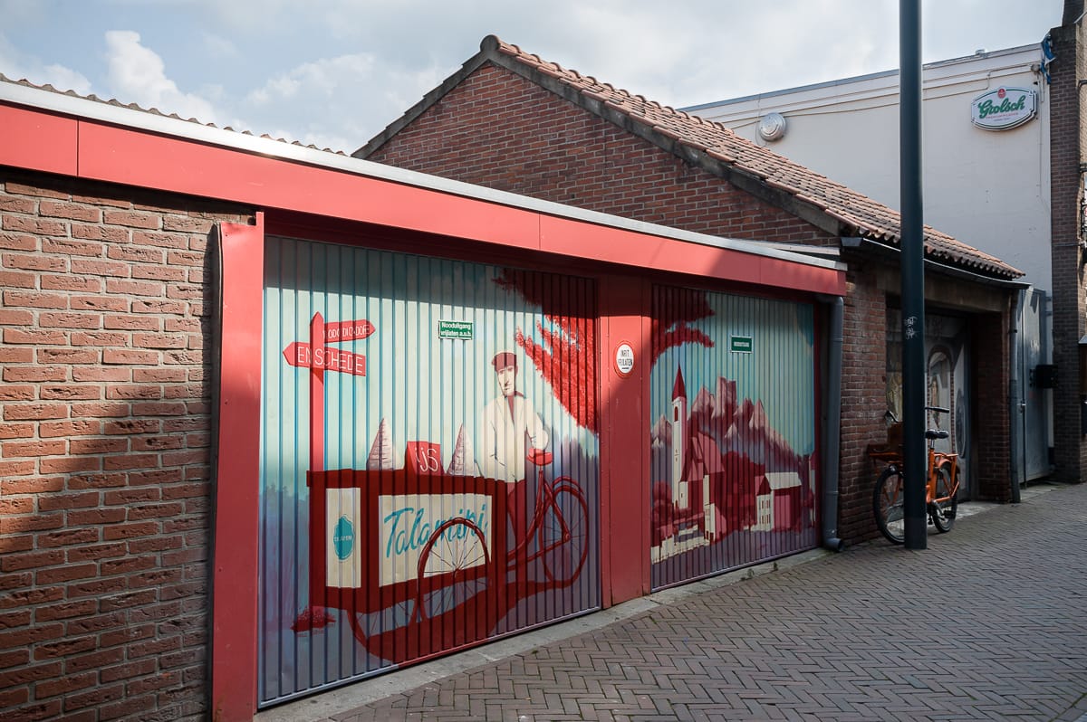 In der Gasse von Enschede zeigen Garagentore ein Wandgemälde, das einen Mann mit Mütze zeigt, der zwischen Bäumen und Häusern ein Oldtimer fährt. Das rot-blaue Kunstwerk ziert ein Backsteingebäude mit Ziegeldach, während unter dem Radeloos-Straßenschild ein Fahrrad steht.