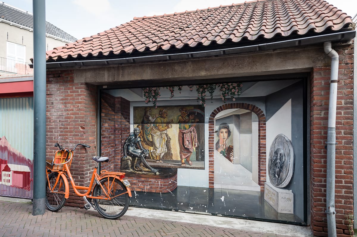 Ein Wandgemälde an einer Backsteinmauer in Enschede zeigt eine klassische Szene mit Figuren in traditionellen Gewändern und einer Büste. Ein orangefarbenes Fahrrad mit einem Korb steht vor dem Kunstwerk, das auf ein Garagentor unter einem roten Ziegeldach gemalt ist, in dieser ruhigen Straße mit einem anderen Gebäude in der Nähe.