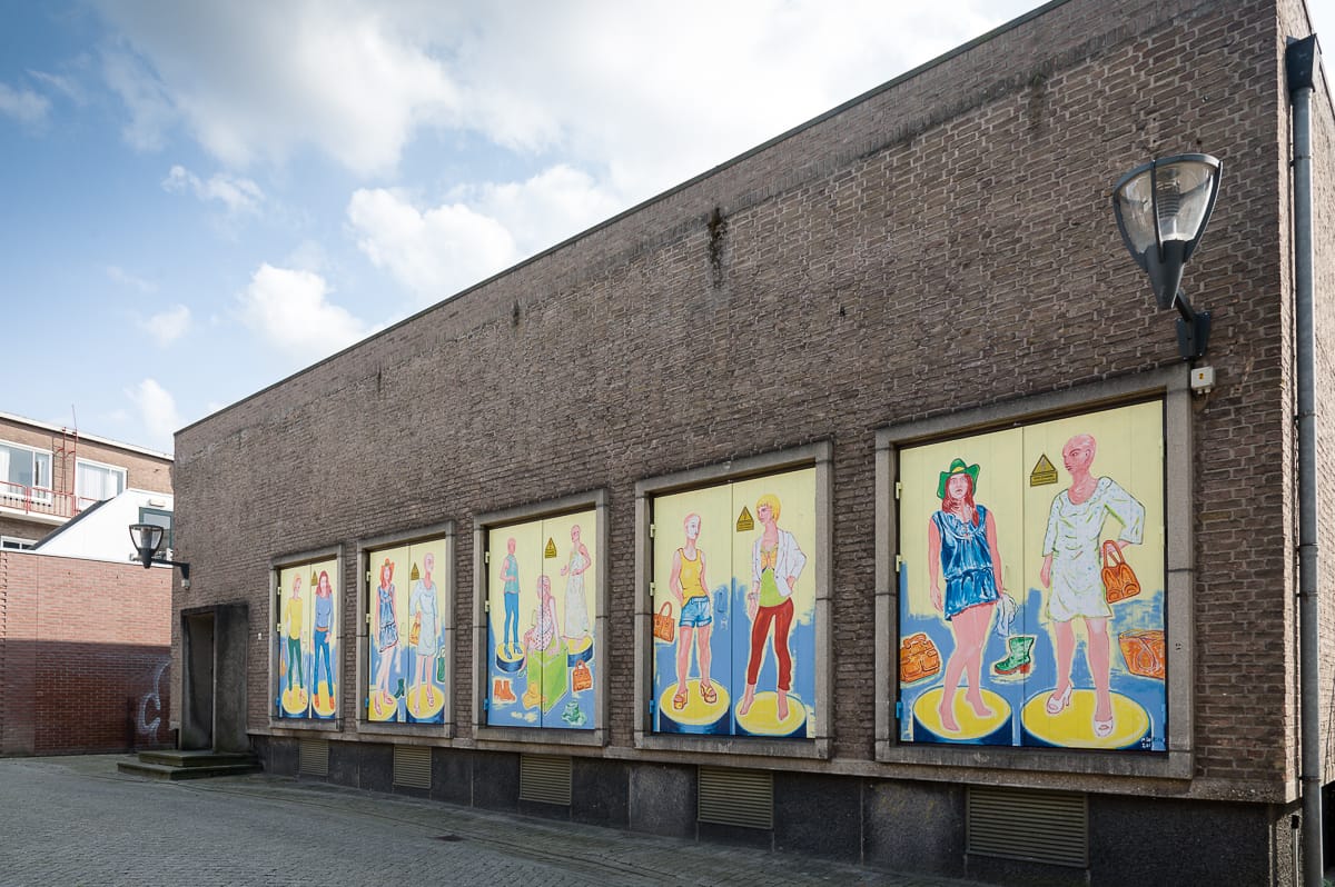 Street Art an einem Backsteingebäude in Enschede besteht aus sechs farbenfrohen, lebensgroßen Tafeln, die stilisierte menschliche Figuren in verschiedenen Posen zeigen. Das Kunstwerk ist lebendig und konzentriert sich auf Mode und Individualität. Auf der rechten Seite ist ein Laternenpfahl zu sehen, der Himmel ist teilweise bewölkt.