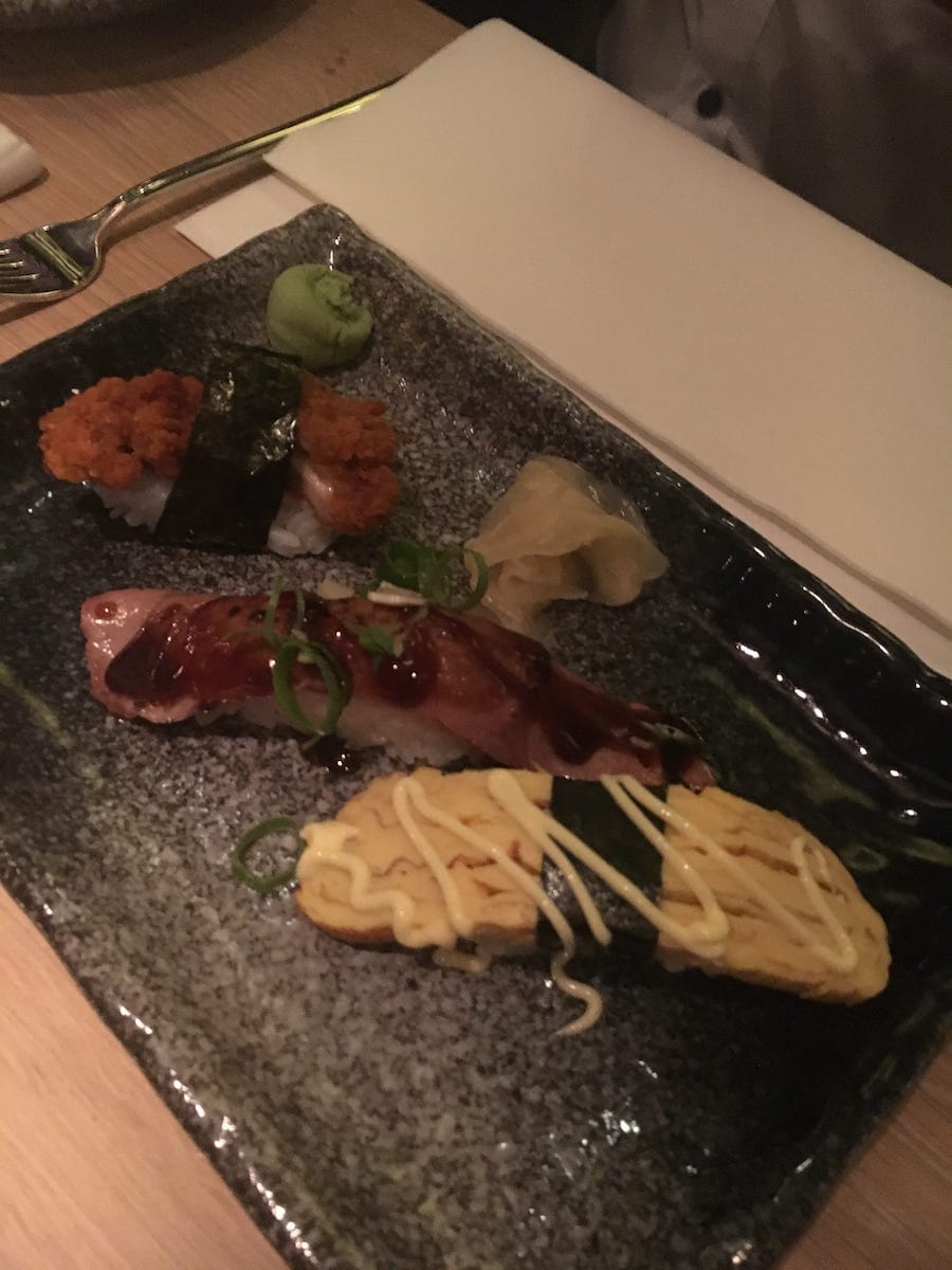 In Enschede liegen auf einem dunklen rechteckigen Teller drei Sushistücke: eines mit frittiertem Belag, ein anderes mit dunkler Soße beträufelt und mit Frühlingszwiebeln garniert und ein drittes mit hellgelbem Ei und Mayonnaise belegt. Wasabi und eingelegter Ingwer liegen neben einer Serviette und einer Gabel.