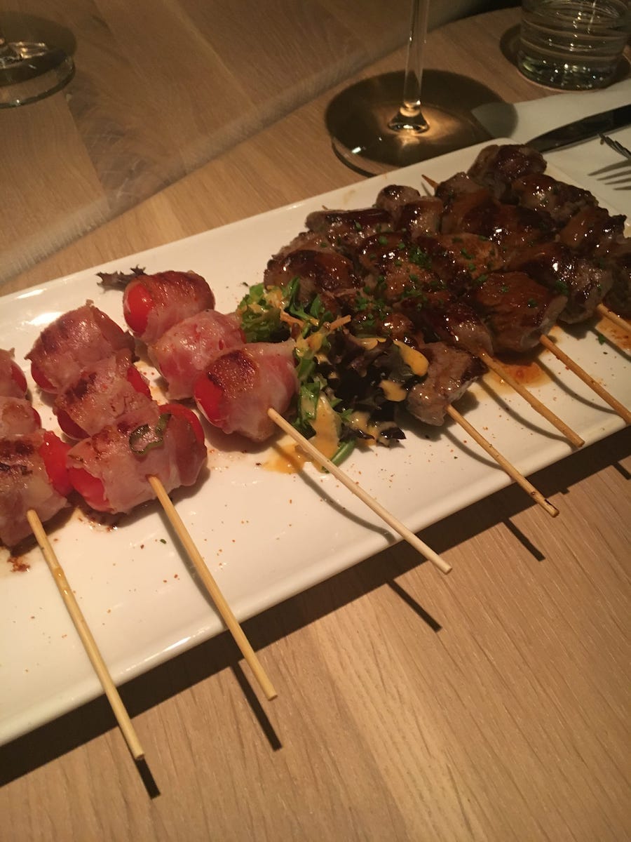 Auf einem rechteckigen weißen Teller auf einem Holztisch in Enschede liegen Spieße mit gegrilltem Fleisch. Links sind Spieße mit Kirschtomaten im Speckmantel zu sehen. Rechts Spieße mit glasiertem Rindfleisch mit Kräutern. Ein Weinglas und ein Wasserglas zieren den Hintergrund.