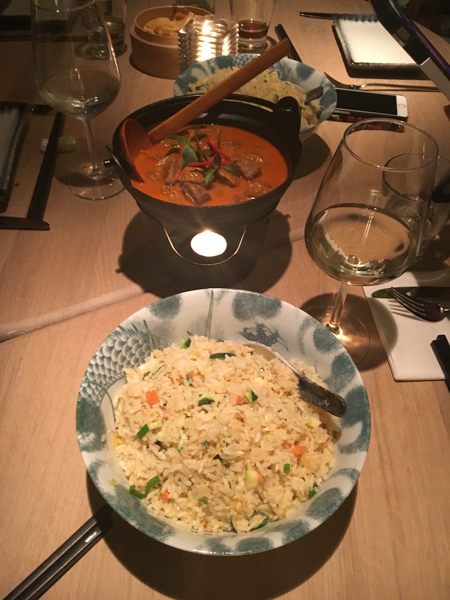 Auf einem Holztisch in Enschede steht eine Schüssel gebratener Reis mit Erbsen und Karotten, ein Topf Orangencurry mit Fleisch und Chilischoten und ein Glas Weißwein. Weitere Gegenstände sind eine brennende Kerze, Essstäbchen, ein Notizbuch und im Hintergrund weitere Teller und Gläser.