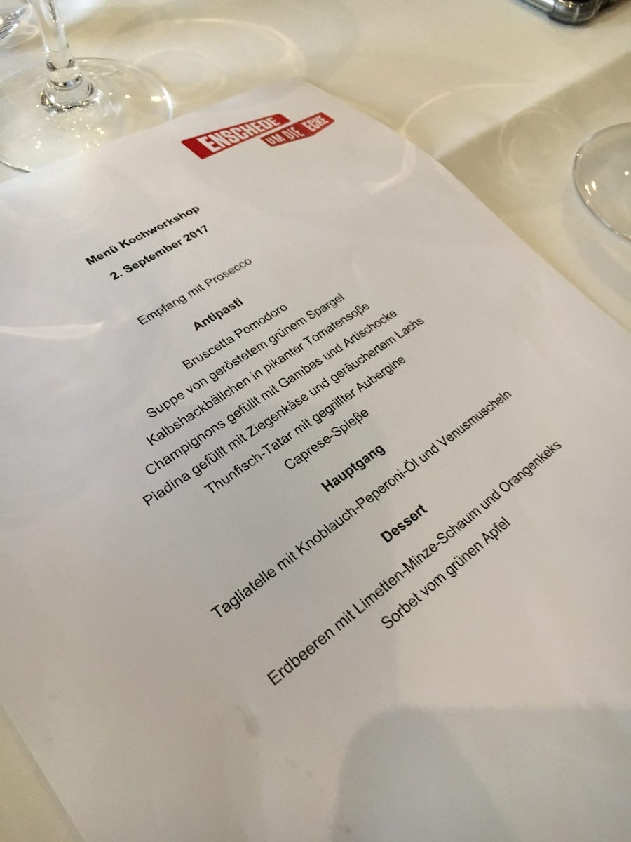 Ein Foto einer gedruckten Speisekarte mit dem Titel „Menü Kochwerkstatt“ aus einem charmanten Restaurant in Enschede vom 2. September 2017. Auf Deutsch sind die Gerichte aufgelistet, darunter Vorspeisen, Hauptgerichte und Desserts. Die Speisekarte liegt auf einer weißen Tischdecke, im Hintergrund sind die Ränder von Weingläsern zu sehen.