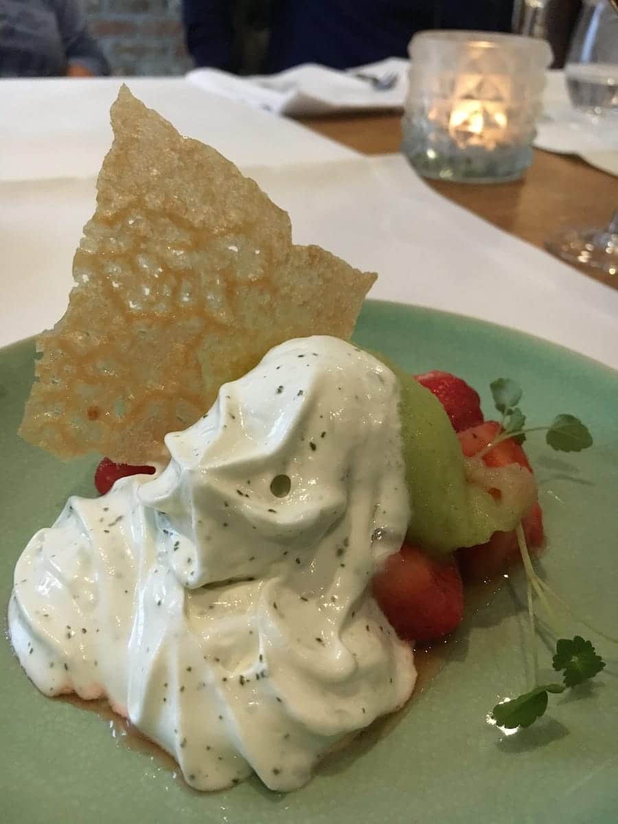 Auf einem grünen Teller in Enschede präsentiert sich ein Dessert mit einem Klecks Schlagsahne und Kräuterstücken. Dazu gibt es geschnittene Erdbeeren und eine Kugel grünes Sorbet. In die Sahne ist eine dünne, knusprige Waffel eingelegt. Im unscharfen Hintergrund sind eine brennende Kerze und Gläser auf einem Holztisch zu sehen.