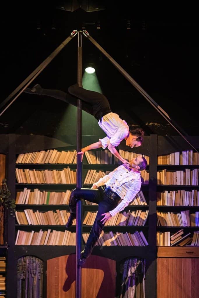 Bénédicte & Simon, Duo Frénésie als leidenschaftliche Romeo & Julia an der Pole im Bookshop, GOP Essen. Sie beugt sich von oben herunter zu ihm, beide mit den Beinen an der Pole festhaltend., liebevoll rechte Hand an seinem Gesicht.