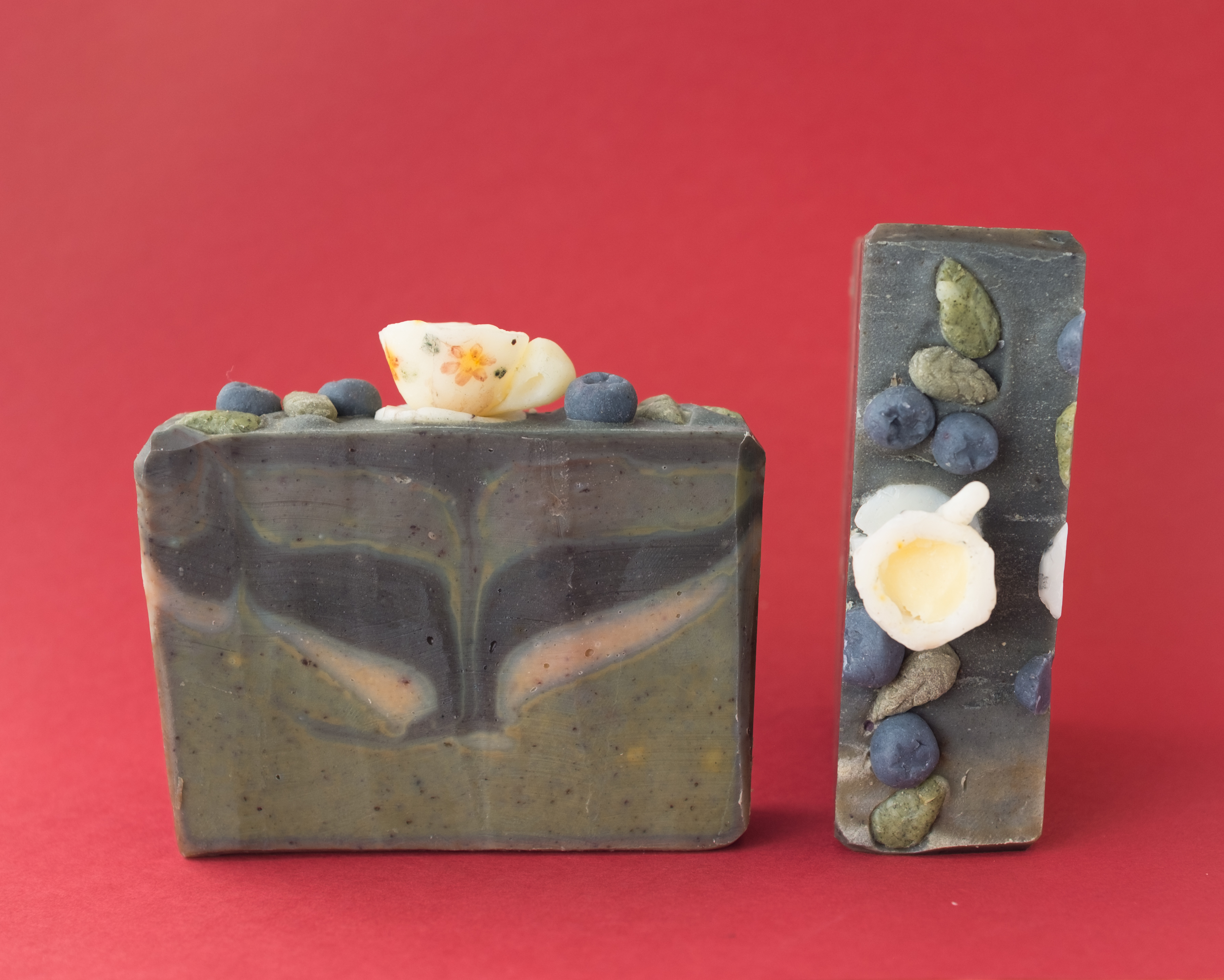 Two decorative Tea Party Soap bars with artistic, colorful layers and embedded details resembling leaves, blueberries, and tiny teacups are displayed against a plain red background.