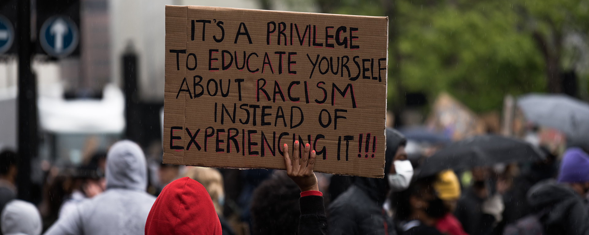 A Black person is seen from behind at a protest, holding a sign. It reads: it's a privilege to educate yourself about racism instead of experiencing it